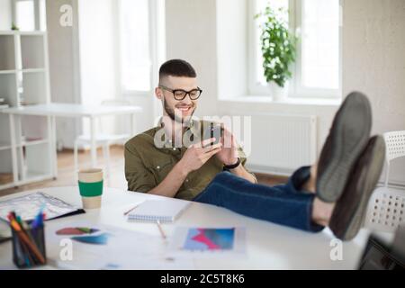 Jeune homme souriant dans des lunettes et chemise joyeusement en utilisant le téléphone portable tout en étant assis sur une chaise avec les jambes sur le bureau au travail. Homme d'affaires joyeux Banque D'Images