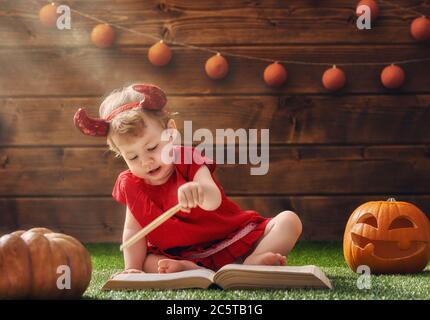 Joyeux Halloween ! Mignon petit diable gai avec baguette magique. Belle enfant bébé fille en costume de diable assis sur l'herbe. Banque D'Images