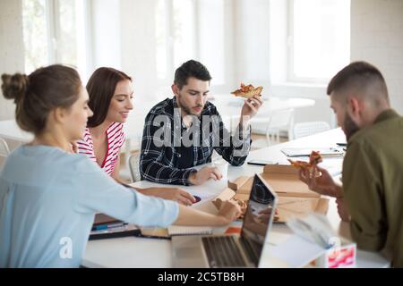 Groupe de jeunes hommes et femmes qui mangent de la pizza ensemble au bureau. Les jeunes créatifs passent du temps au travail Banque D'Images