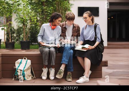 Groupe d'étudiants souriants assis sur le banc et lisant des livres dans la cour de l'université Banque D'Images