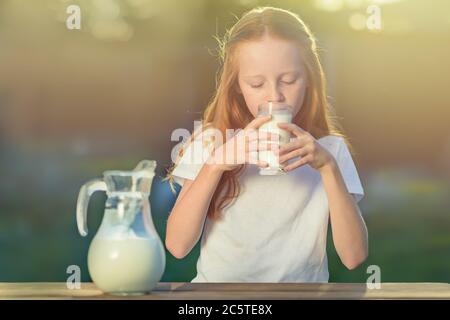 Une fille aux cheveux rouges boit du lait frais d'un verre sur le fond d'une belle matinée ensoleillée dans le village. Un pot de lait de chèvre repose sur un bois Banque D'Images