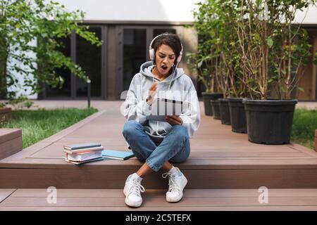 Une jeune femme a choqué les écouteurs assis et regardant avec étonnement la tablette tout en passant du temps dans la cour de l'université Banque D'Images