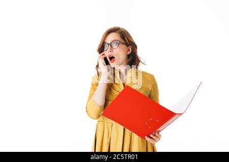 Portrait de jeune femme choquée dans des lunettes debout avec le téléphone portable et ouvrir le dossier rouge dans les mains tout en regardant de côté sur fond blanc Banque D'Images