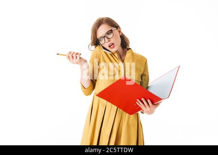 Portrait d'une femme occupée à lunettes parlant sur son téléphone portable tout en regardant avec colère de côté avec dossier rouge ouvert et crayon à la main sur fond blanc Banque D'Images