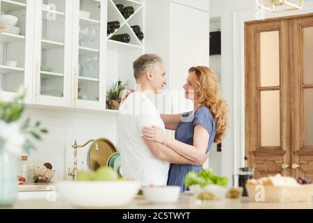 Portrait de couple d'âge mûr aimant embrassant et regardant l'un l'autre dans l'intérieur blanc de la cuisine à la maison, espace de copie Banque D'Images