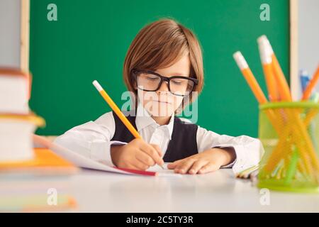 Scolarité. Un écolier intelligent avec des lunettes écrit une affectation d'enseignant dans un bloc-notes à une table dans la salle de classe. Banque D'Images