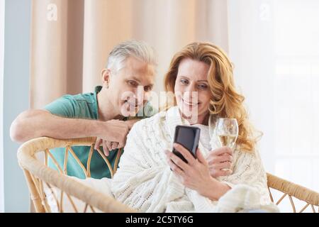 Portrait aux tons chauds d'un couple élégant et mûr regardant l'écran du smartphone tout en se relaxant à la maison, espace de copie Banque D'Images