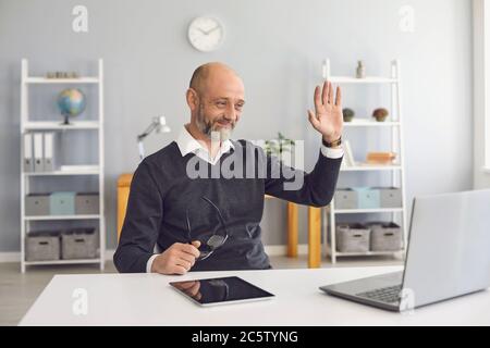 Un homme senor moderne heureux accueille avec des mains tenir un ordinateur portable de vidéoconférence, assis à la table du bureau à domicile. Banque D'Images