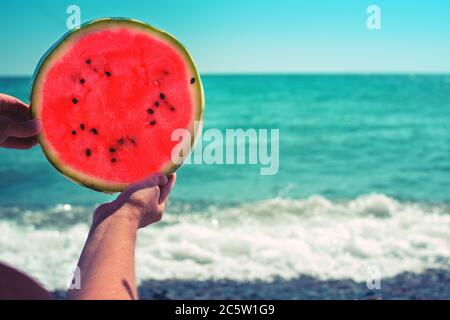 Pastèque ronde, tranche juteuse sous forme de soleil, entre les mains d'une femme contre la mer, sous les rayons du soleil. Régime de fruits tropicaux. Banque D'Images