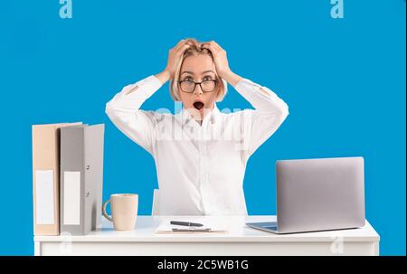 Une secrétaire mature tient sa tête en choc sur son lieu de travail avec un ordinateur portable, fond bleu Banque D'Images