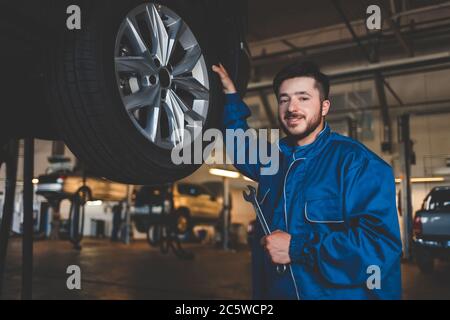 Mécanicien automatique avec des supports de clé près d'une voiture sur un ascenseur. Le réparateur de voiture dans un garage tient sa main sur une roue de voiture Banque D'Images