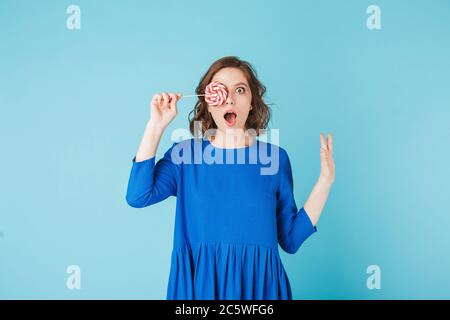 Portrait de la jolie dame en robe debout et couvrant son visage avec des bonbons à la lylipop tout en regardant avec étonnement dans l'appareil photo sur fond bleu Banque D'Images