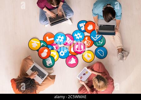 Vue de dessus de quatre jeunes gens amicaux en tenue décontractée assis par des papiers multicolores avec des icônes sur le sol et utilisant des gadgets mobiles Banque D'Images