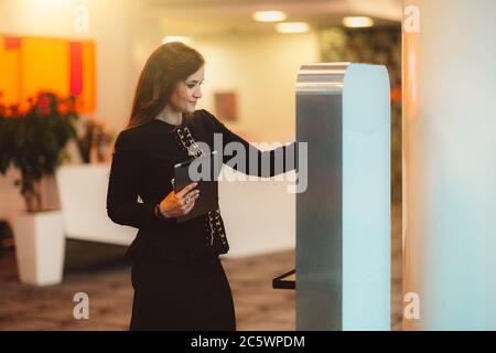Une femme d'entrepreneur cacacasien, charmante et majestueuse, en costume noir formel, avec une tablette numérique dans une main, interagit avec un terminal électronique Banque D'Images