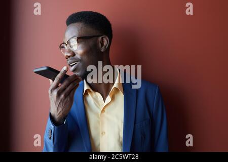 Portrait d'un homme afro-américain élégant enregistrant un message vocal via un smartphone et portant des lunettes tout en posant sur un arrière-plan marron, espace de copie Banque D'Images