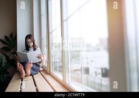 Une jeune fille adorable lit le livre à la maison tout en étant assise sur le rebord de la fenêtre Banque D'Images