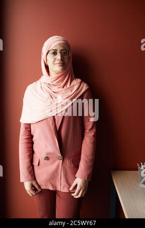 Portrait vertical d'une femme d'affaires réussie portant un foulard regardant l'appareil photo tout en se posant en toute confiance contre le mur rouge au bureau Banque D'Images
