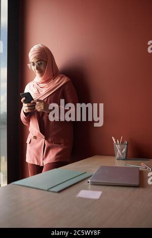 Portrait vertical de la jeune femme d'affaires musulmane utilisant un smartphone tout en se tenant contre le mur rouge dans le bureau, espace de copie Banque D'Images