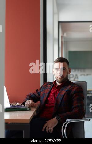 Portrait vertical d'un homme d'affaires barbu élégant regardant l'appareil photo tout en étant assis sur le lieu de travail et en posant avec confiance contre le mur rouge au bureau, copie Banque D'Images