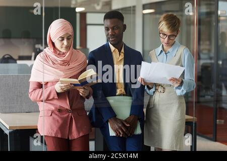 Portrait à la taille de l'équipe professionnelle multiethnique moderne composée de trois personnes qui tiennent des documents tout en se tenant derrière un mur de verre au bureau Banque D'Images