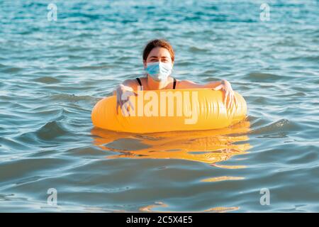 Protection contre les virus et la distance sociale. Une femme dans un masque médical nage dans la mer avec un cercle gonflable. Copier l'espace. Banque D'Images