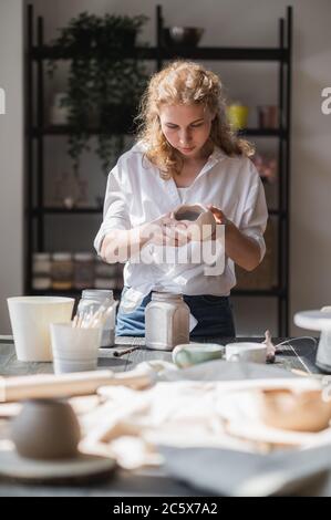 La femelle potter assis et agite la peinture avec un pinceau une tasse sur la table. Femme en céramique. Travail de poterie, compétences artisanales et créatives. Banque D'Images