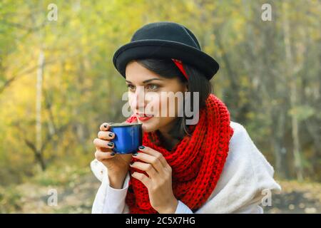 Belle jeune femme en manteau blanc, foulard rouge crochet et chapeau noir tient une tasse dans ses mains. Fête du thé dans la nature Banque D'Images
