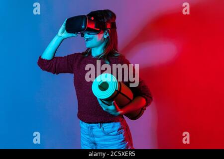 Bonne fille avec un casque VR lunettes tenant un tapis de yoga roulé, regardant des cours de fitness et souriant excité par les activités sportives en réalité virtuelle, wor Banque D'Images