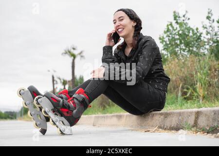 Une femme heureuse en train de faire une pause pour le patinage à roulettes, assise dans la rue et utilisant un téléphone portable. Jolie, Urban Girl parlant au téléphone, portant des patins à roulettes. Fille moderne dans la ville . Photo de haute qualité Banque D'Images