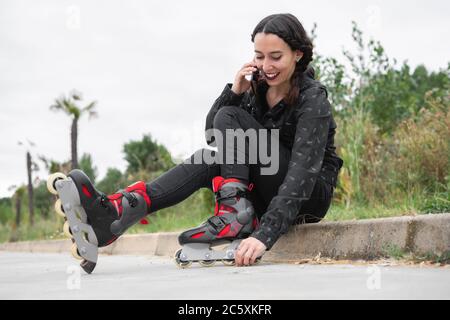 Une femme heureuse en train de faire une pause pour le patinage à roulettes, assise dans la rue et utilisant un téléphone portable. Jolie, Urban Girl parlant au téléphone, portant des patins à roulettes. Fille moderne dans la ville . Photo de haute qualité Banque D'Images