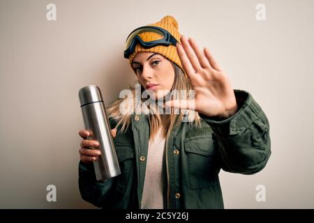 Jeune belle femme de skieur de brunette portant des lunettes de ski buvant thermo avec café à main ouverte faisant signe d'arrêt avec la sérieuse et confiante expressi Banque D'Images