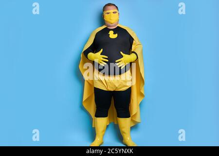 portrait de graisse de nettoyage caucasien super-héros touchant son estomac, homme de héros en costume jaune posant à l'appareil photo Banque D'Images