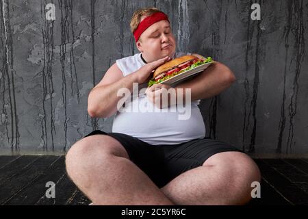 fat l'homme caucasien tient un sandwich comme un enfant, le mâle est assis sur le sol et il est impatient de le manger, un concept malsain de manger Banque D'Images