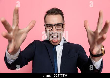 modèle élégant et heureux en costume portant des lunettes, hurlant et riant, tenant les mains vers le haut et debout sur fond rose Banque D'Images