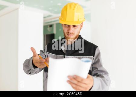 Un jeune ingénieur sérieux dans les vêtements de travail et le casque jaune cependant Banque D'Images