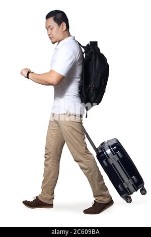 Homme asiatique voyageur marchant vers l'avant traînez ses bagages de valise tout en regardant sa montre, portant une chemise décontractée et un sac à dos, portrait pleine longueur isola Banque D'Images