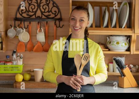 Jolie jeune femme au foyer en tablier noir pose dans la cuisine de maison de campagne confortable Banque D'Images