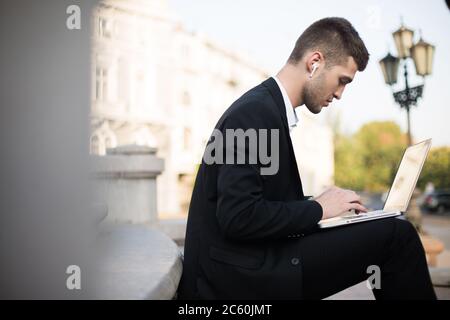 Jeune homme d'affaires élégant en costume classique avec écouteurs sans fil travaillant intelligemment sur un ordinateur portable extérieur Banque D'Images