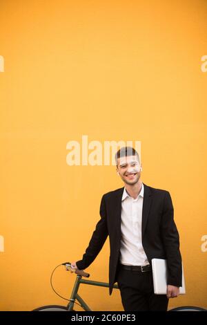 Jeune homme joyeux en costume noir classique et chemise blanche avec écouteurs sans fil tenant un ordinateur portable à la main tout en regardant joyeusement dans l'appareil photo avec vélo Banque D'Images