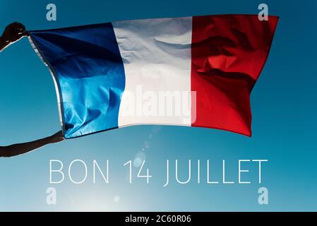 Homme portant un drapeau français agitant sur le vent et le texte bon 14 juillet, heureux 14 juillet, la journée nationale de la France écrite en français, contre le blu Banque D'Images