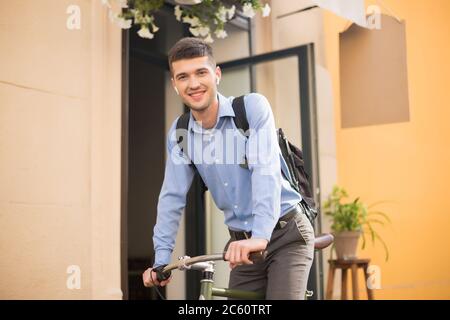 Jeune homme beau en chemise bleue avec écouteurs sans fil et sac à dos à vélo tout en regardant avec plaisir dans l'appareil photo extérieur Banque D'Images