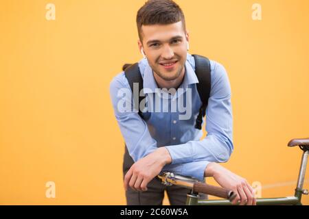 Jeune homme souriant en chemise bleue et sac à dos noir avec écouteurs sans fil, incliné sur vélo tout en regardant avec plaisir dans l'appareil photo sur fond orange Banque D'Images
