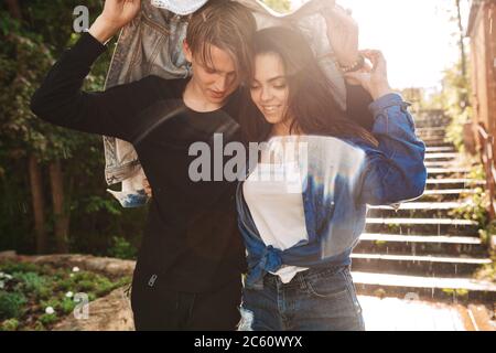 Portrait d'un jeune couple heureux qui se couvre avec une veste en denim tout en se retrouvant sous la pluie dans le parc Banque D'Images