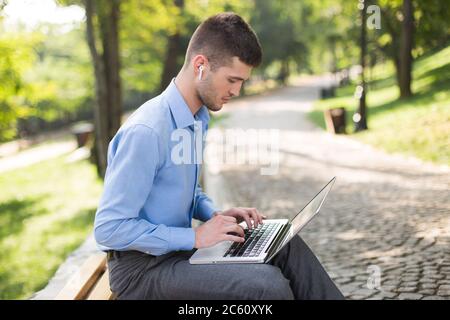 Jeune homme beau en chemise bleue avec écouteurs sans fil assis sur un banc, travaillant intelligemment sur un ordinateur portable tout en passant du temps dans le parc Banque D'Images