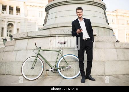 Jeune homme d'affaires souriant, costume noir classique et chemise blanche, avec écouteurs sans fil tenant le téléphone portable à la main tout en regardant de rêve dans l'appareil photo Banque D'Images