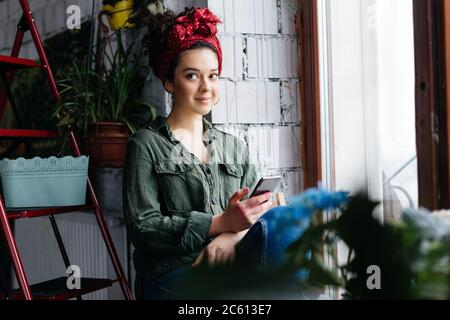 Jeune femme souriante aux cheveux maures sombres, assise près d'une échelle avec des fleurs tenant un téléphone portable à la main tout en regardant de côté dans un confortable et moderne Banque D'Images