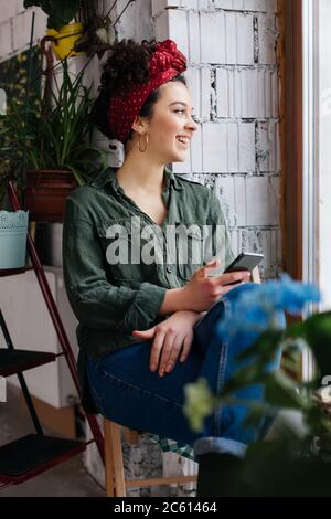 Jeune femme joyeuse aux cheveux fclés foncés assise près de l'échelle avec des fleurs tenant le téléphone portable en main tout en regardant avec joie de côté dans un atelier moderne et confortable Banque D'Images