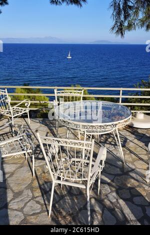 Table de pique-nique et chaises sous les pins dans la cour sur la côte de la mer Égée au coucher du soleil, yacht blanc à voile sur fond. Lumière chaude du soir. A Banque D'Images