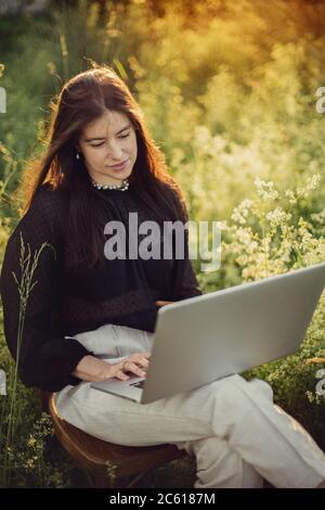 Nouveau concept de bureau. Élégante fille à la mode travaillant sur un ordinateur portable et assise sur une chaise rustique dans la lumière chaude du coucher de soleil dans le champ d'été. Travailler à distance avec soci Banque D'Images