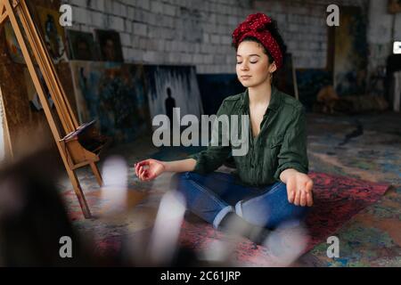 Jeune belle femme avec les yeux fermés assis sur le tapis de yoga dans la pose de lotus et méditant tout en passant du temps dans l'atelier confortable Banque D'Images